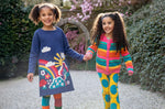 2 children holding hands in a garden setting. One child is wearing the Frugi Kari navy rainbow dress, the other child is wearing the Frugi bight rainbow striped cardigan and super dot trousers available at Babipur.