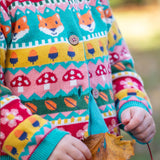 A child leaning on a tree and wearing the Frugi kids robyn fairisle cardigan in fox. A 100% GOTS organic cotton cardigan with a bright woodland fairisle design and button fasteners