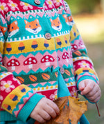 A child leaning on a tree and wearing the Frugi kids robyn fairisle cardigan in fox. A 100% GOTS organic cotton cardigan with a bright woodland fairisle design and button fasteners