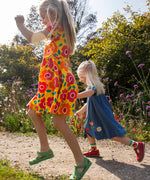 Child wearing Frugi's yellow spring skater dress with repeating bright floral designs and skipping on a path next to another child