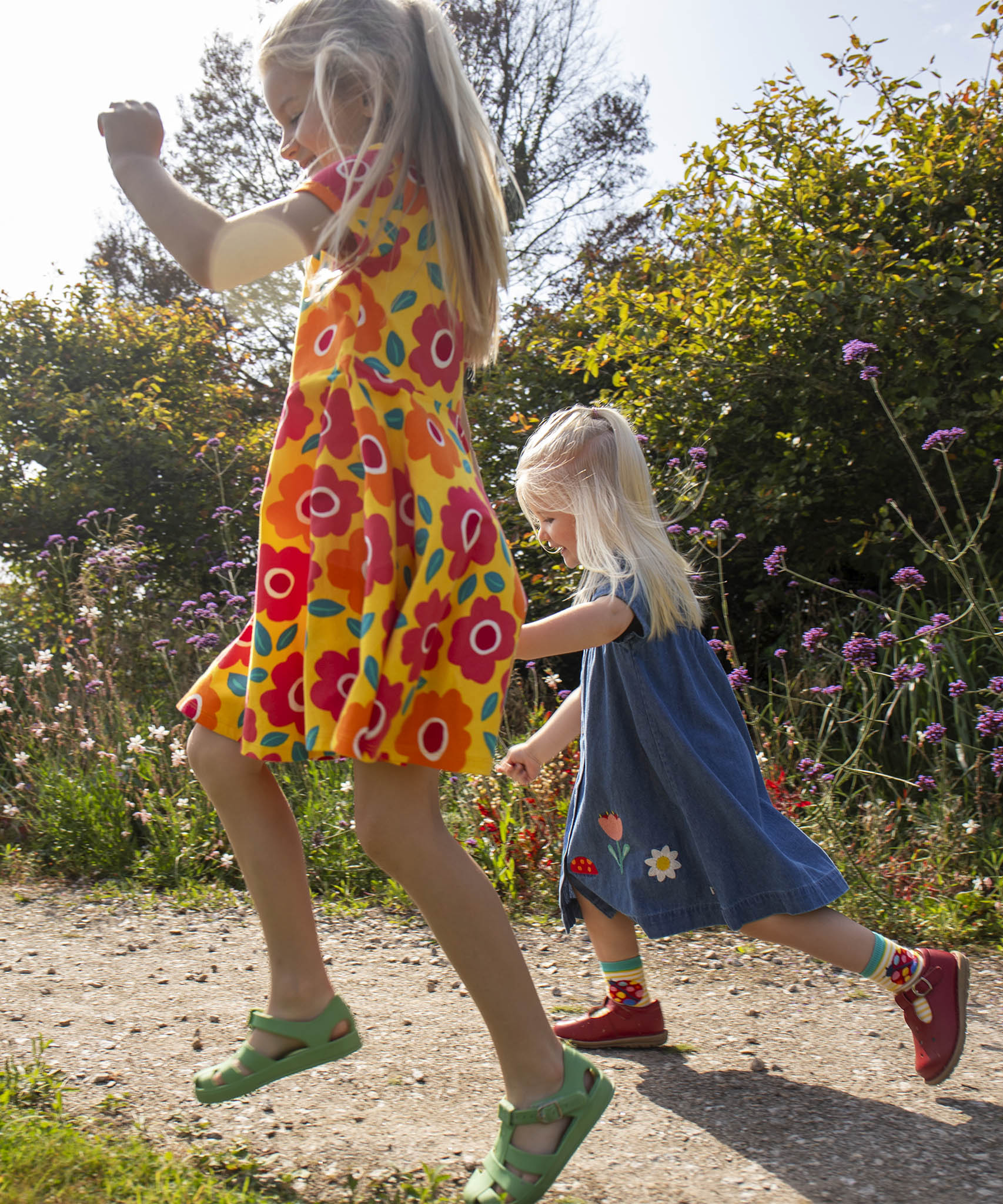 Child wearing Frugi's yellow spring skater dress with repeating bright floral designs and skipping on a path next to another child