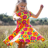 Child wearing Frugi's yellow spring skater dress with repeating bright floral designs. Standing in a field with trees in the background.