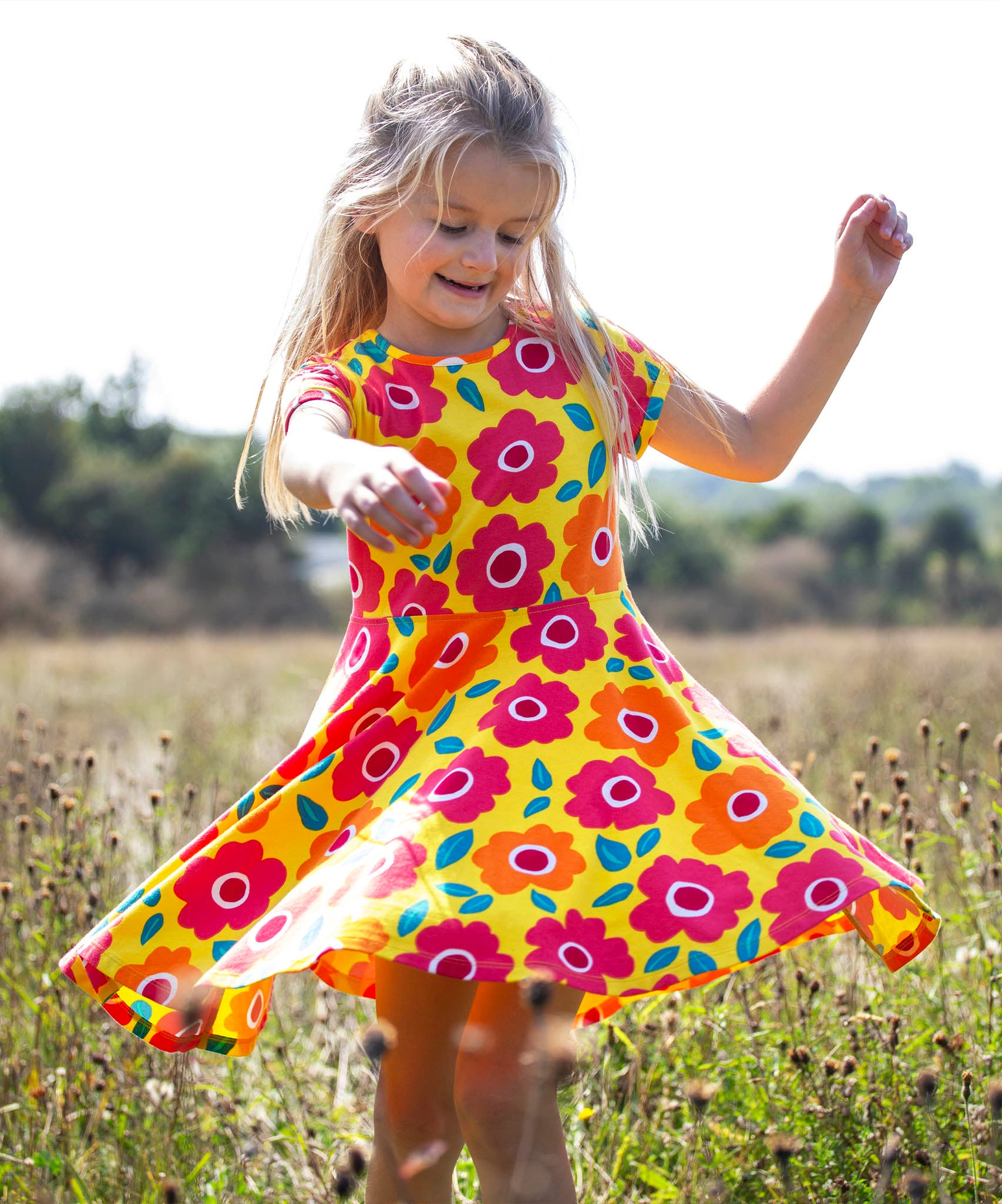 Child wearing Frugi's yellow spring skater dress with repeating bright floral designs. Standing in a field with trees in the background.