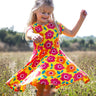 Child wearing Frugi's yellow spring skater dress with repeating bright floral designs. Standing in a field with trees in the background.