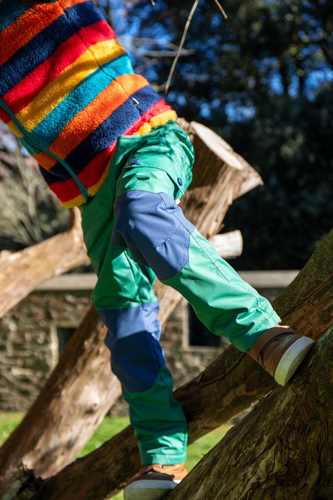 A child standing on a tree and wearing the Frugi Wilderness green trousers with navy blue coloured knee patches available at Babipur