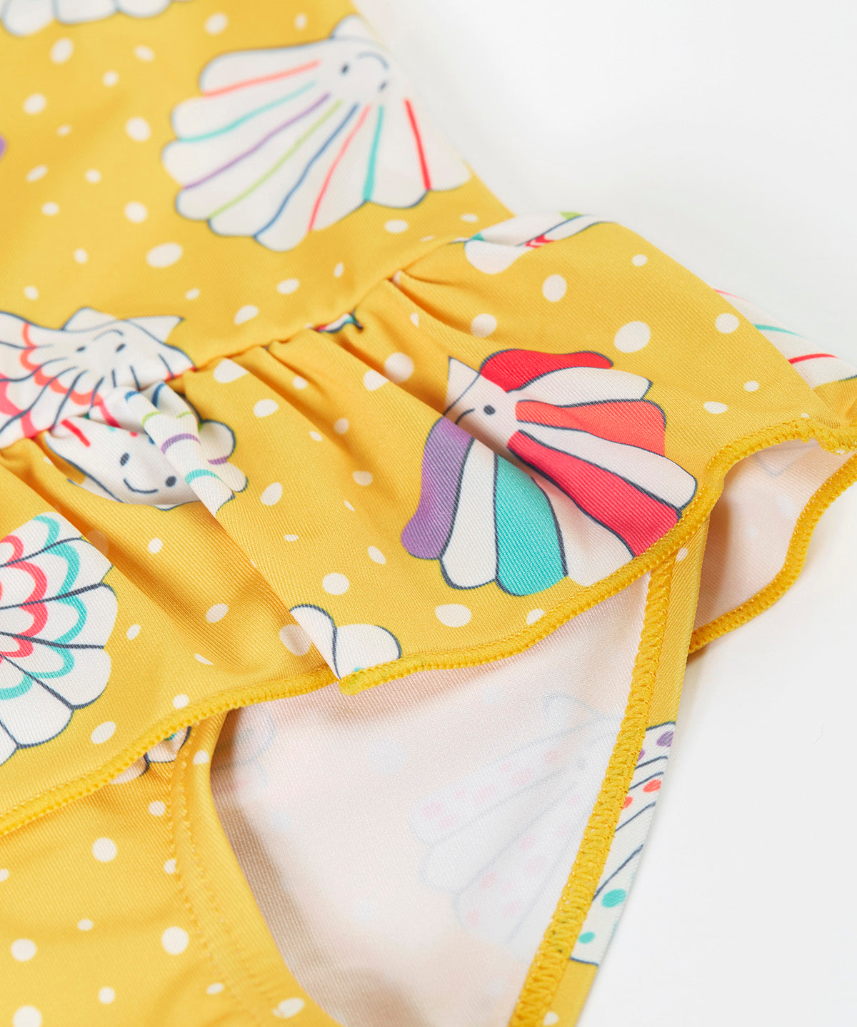 A close up of the Frugi yellow swim suit laid flat showing the repeating white shell design and frill detail on the waist.