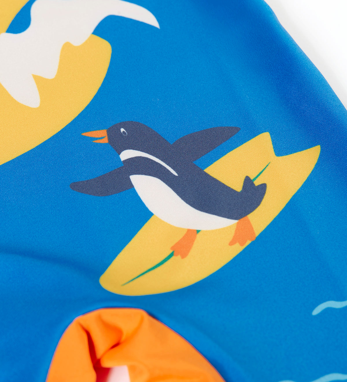 A close up of the surfing penguin on the blue Frugi sun safe all in one swimwear suit