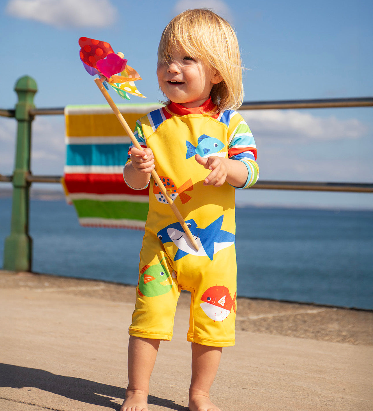 Child smiling and standing on a concret surface with the sea in the background. Holding a toy windmill and wearing a Frugi yellow all in one sun safe swimwear suit with different sea creatures