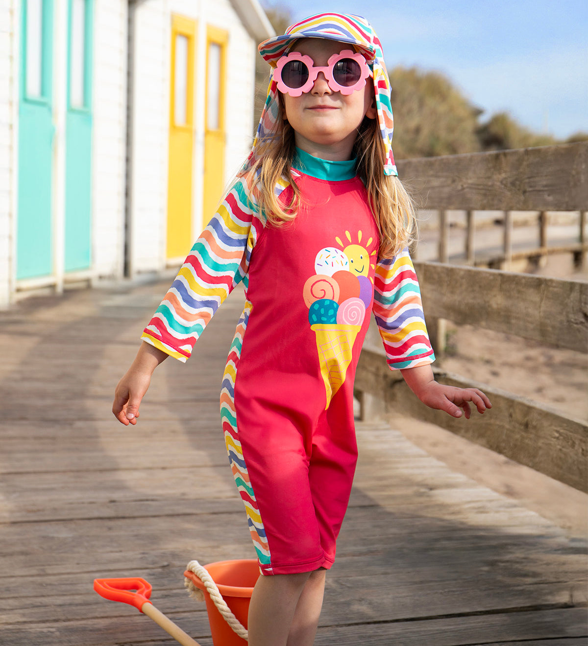 Child wearing pink sunglasses and standing on a wooden path with beach huts in the background. Wearing the pink Frugi  sun safe swimwear suit with pastel coloured striped sleeves and a ice cream on the front. 