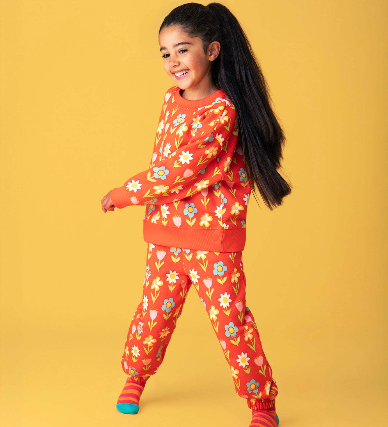 A child smiling and standing in front of a light orange background. Wearing a red Frugi sweater and matching joggers with a repeating flower print.