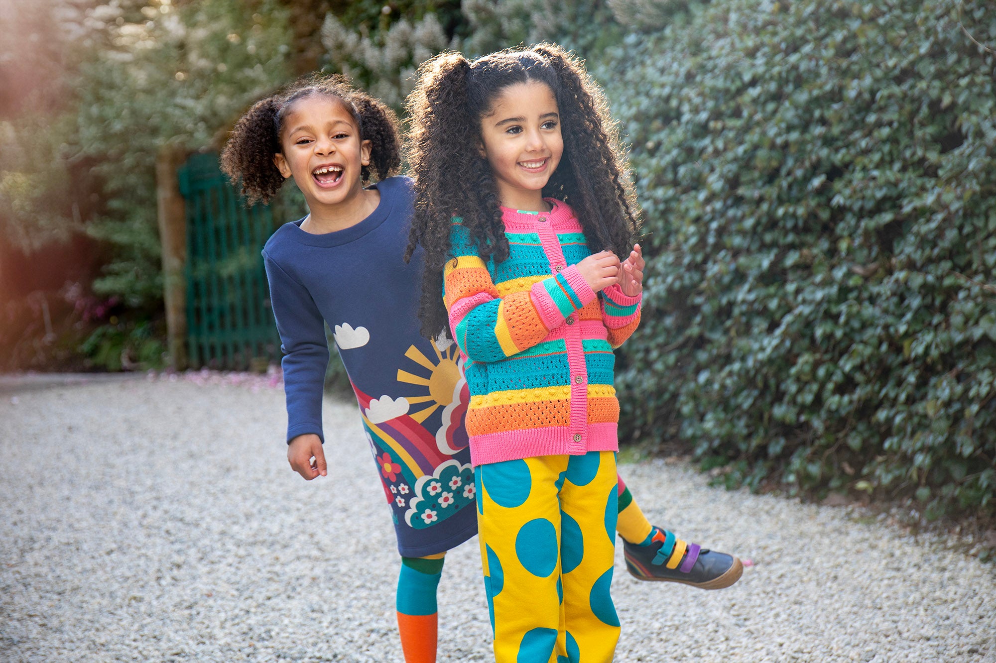 A child wearing the Frugi Kari navy rainbow dress and playing behind  another child wearing the Frugi bight rainbow striped cardigan and super dot trousers available at Babipur.
