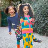 A child wearing the Frugi Kari navy rainbow dress and playing behind  another child wearing the Frugi bight rainbow striped cardigan and super dot trousers available at Babipur.