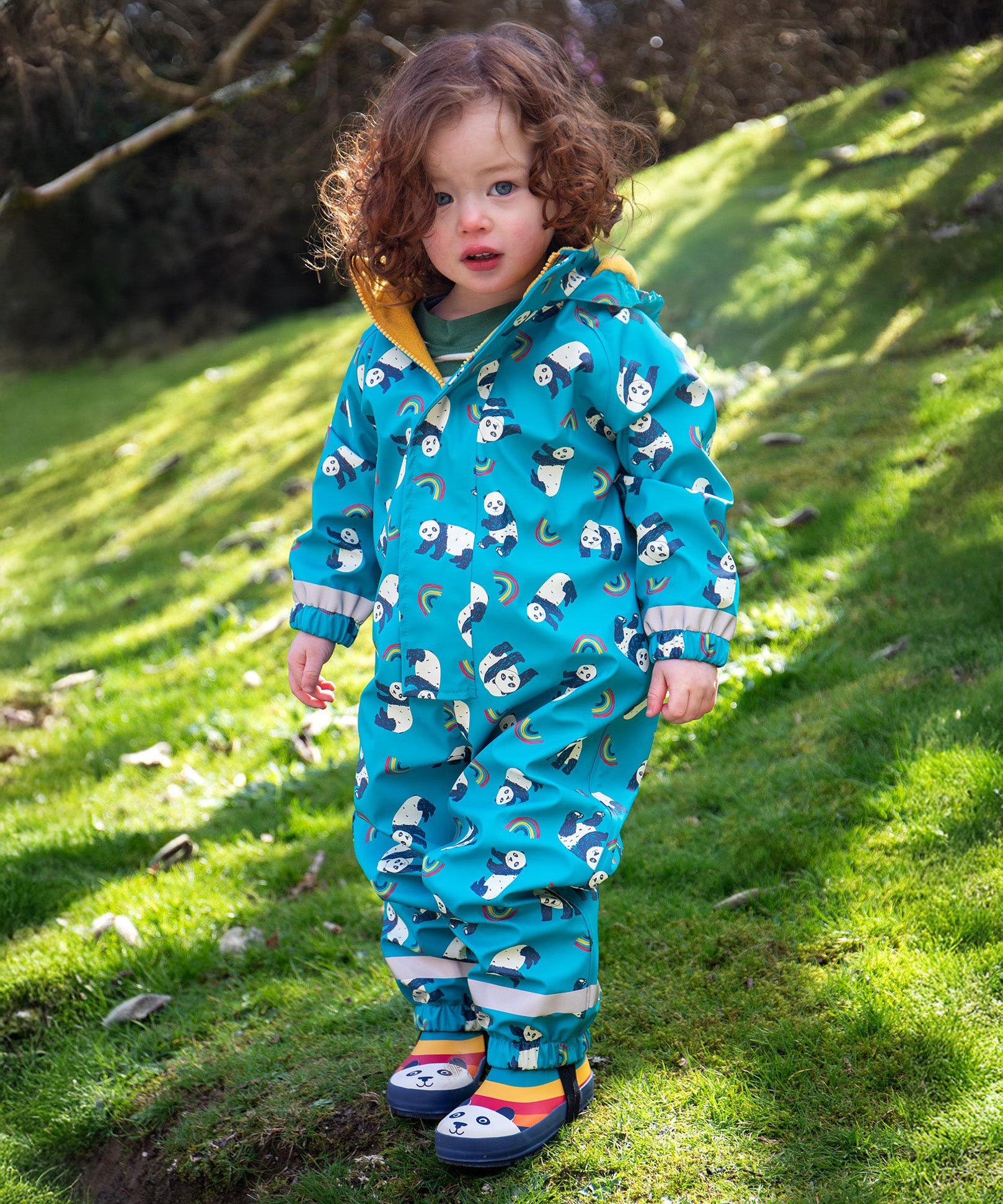 A toddler wearing the Frugi puddle buster blue panda play waterproof all in one suit with the panda wellingtons. 