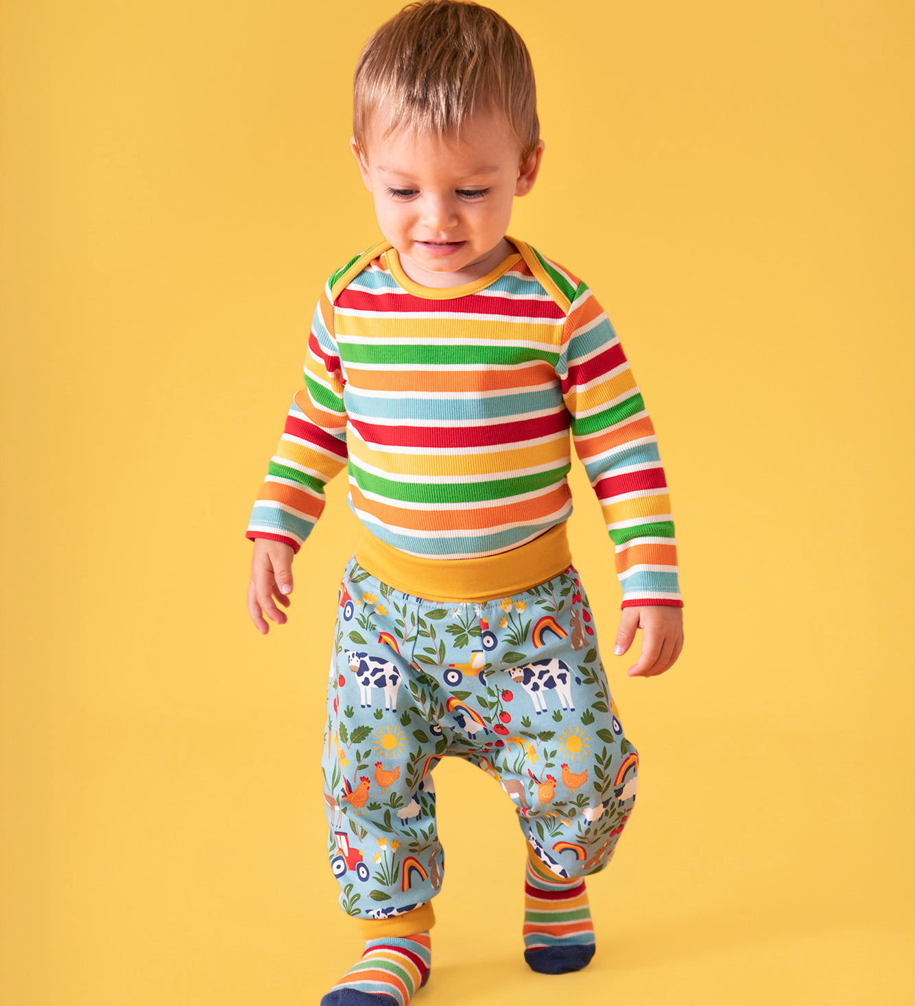 Toddler standing against a light orange background, wearing a bold striped long sleeve top, socks and the repeated farm inspired print parsnip pants