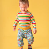 Toddler standing against a light orange background, wearing a bold striped long sleeve top, socks and the repeated farm inspired print parsnip pants