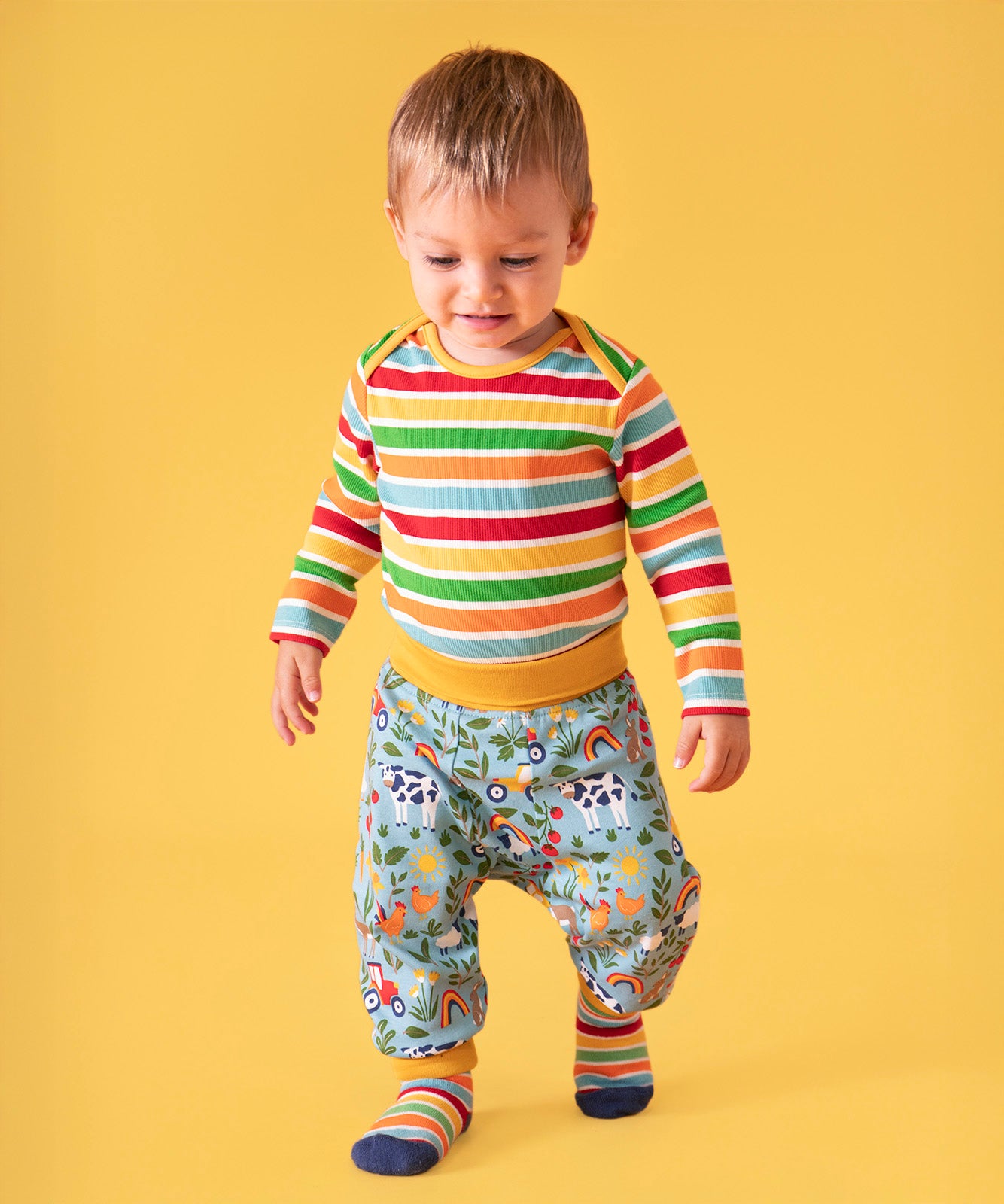 Toddler standing against a light orange background, wearing a bold striped long sleeve top, socks and the repeated farm inspired print parsnip pants