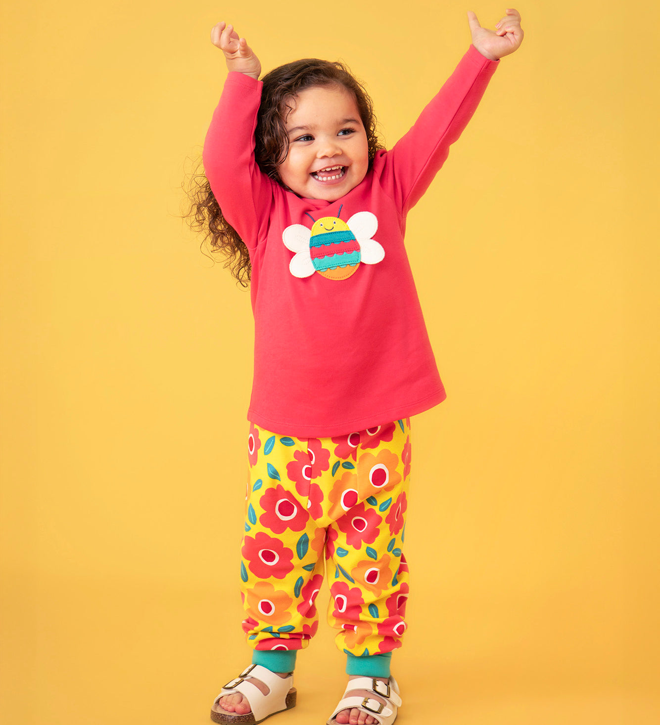 Toddler standing against a light orange background wearing a pink long sleeve top with a bee print and the Frugi floral yellow pants