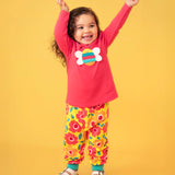 Toddler standing against a light orange background wearing a pink long sleeve top with a bee print and the Frugi floral yellow pants