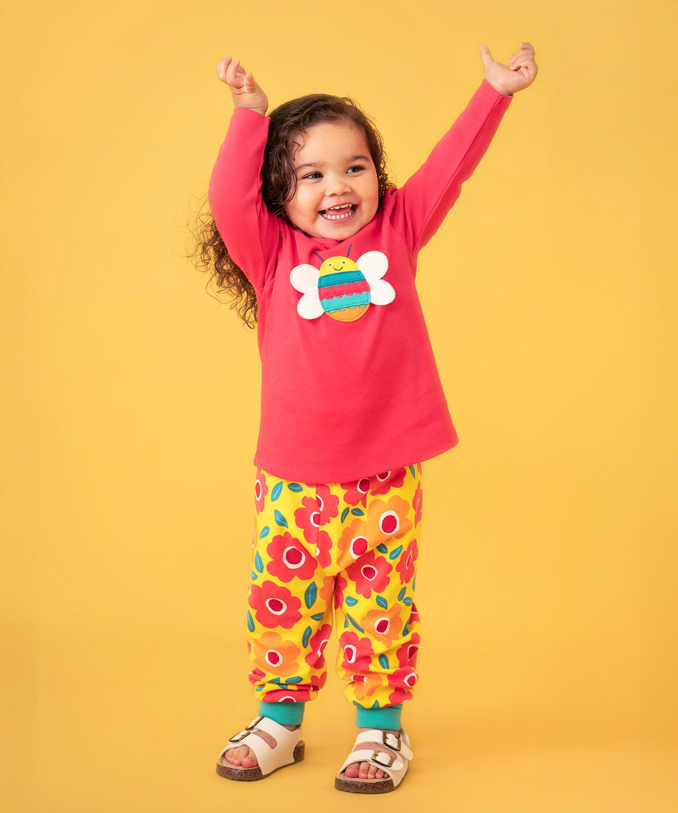 Toddler standing against a light orange background wearing a pink long sleeve top with a bee print and the Frugi floral yellow pants
