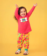Toddler standing against a light orange background wearing a pink long sleeve top with a bee print and the Frugi floral yellow pants