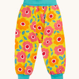 Frugi organic cotton parsnip pants laid flat. In bright yellow with a turquoise waist band and leg cuffs. Has a repeating floral print. 