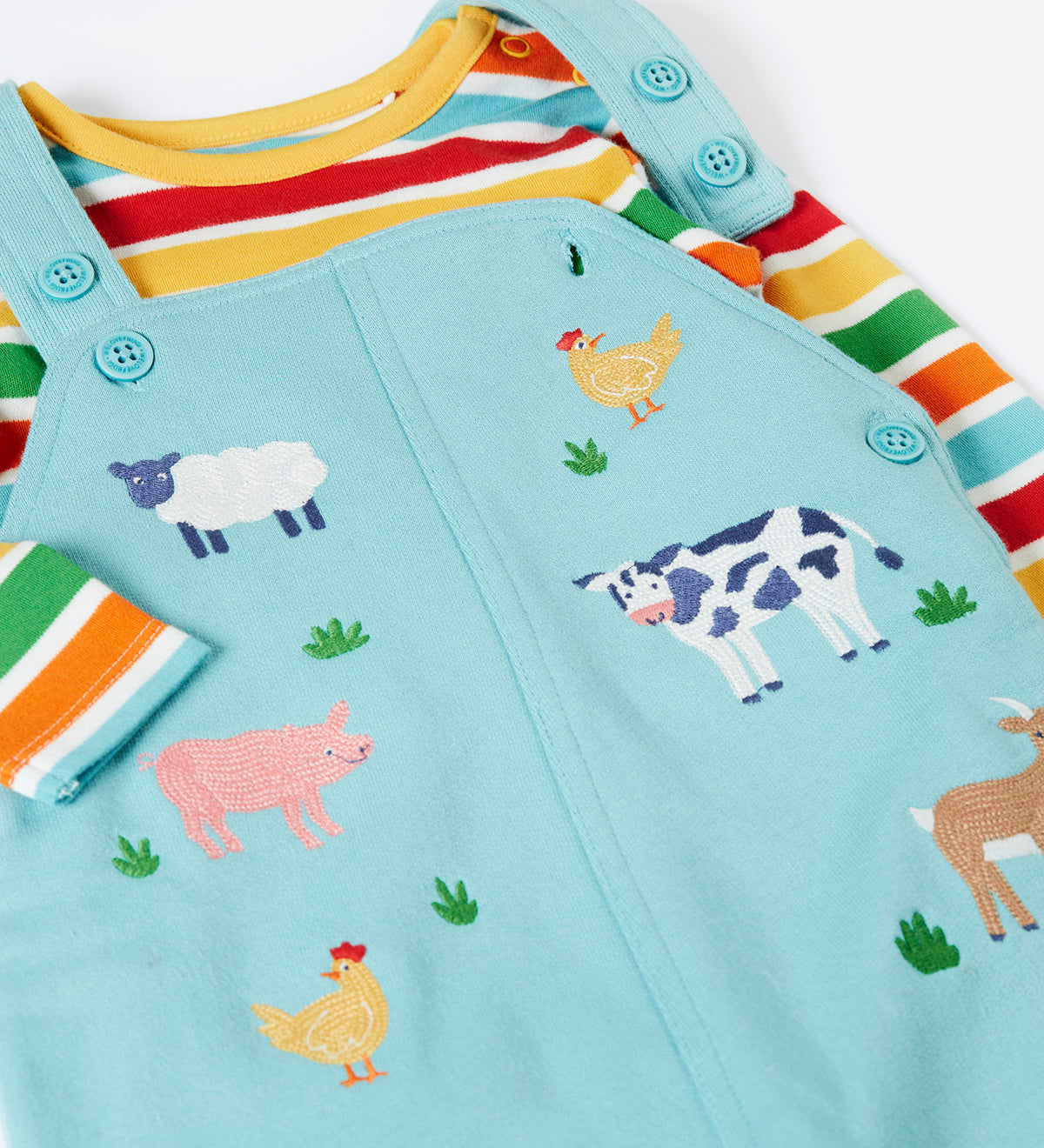 A close up of the Frugi light blue organic cotton dungaree with farm animals on the front laid flat with a rainbow striped long sleeve top underneath as an outfit
