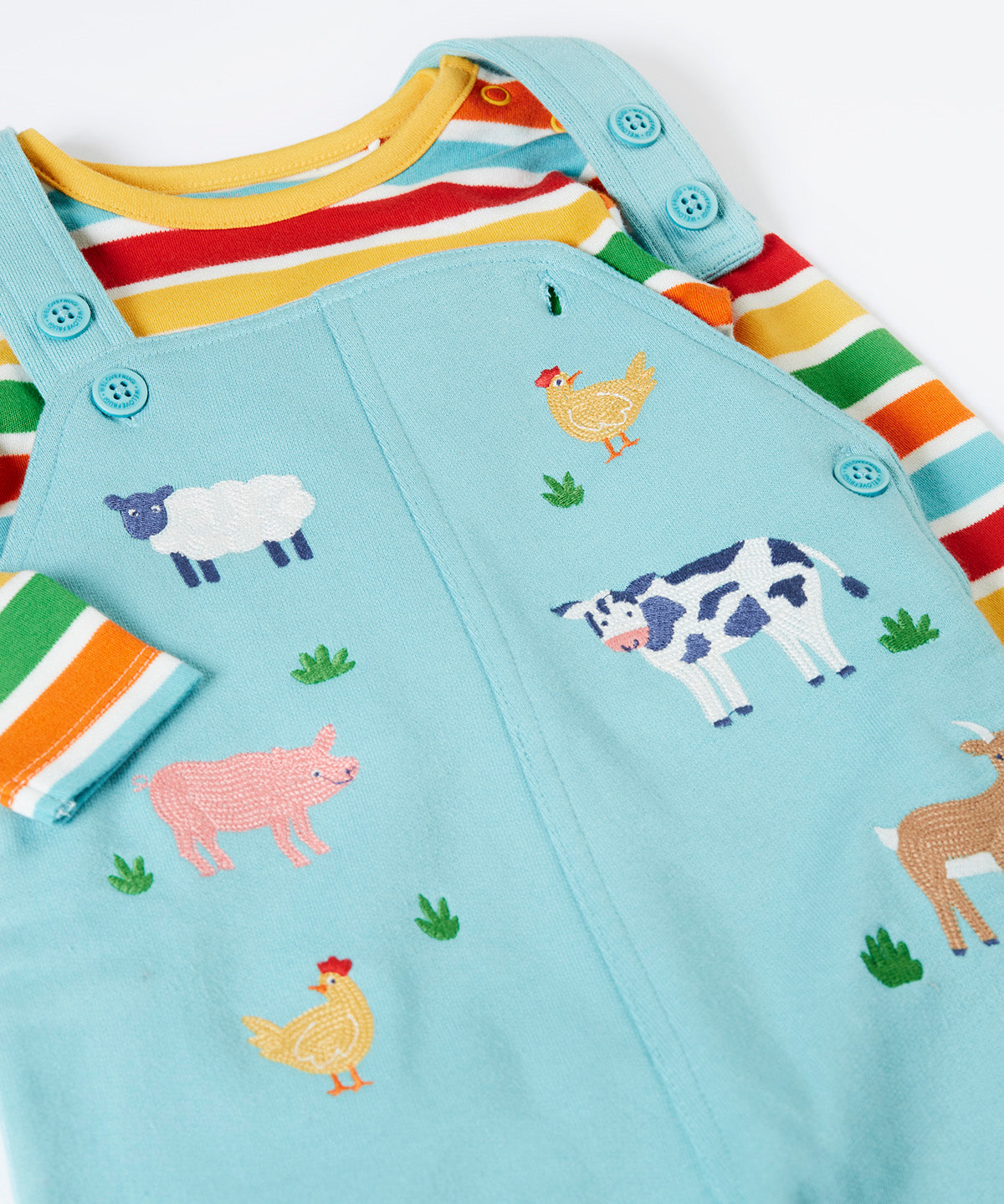 A close up of the Frugi light blue organic cotton dungaree with farm animals on the front laid flat with a rainbow striped long sleeve top underneath as an outfit