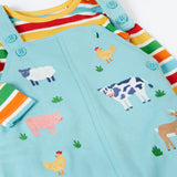 A close up of the Frugi light blue organic cotton dungaree with farm animals on the front laid flat with a rainbow striped long sleeve top underneath as an outfit
