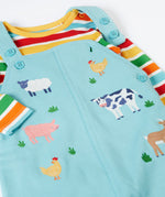 A close up of the Frugi light blue organic cotton dungaree with farm animals on the front laid flat with a rainbow striped long sleeve top underneath as an outfit