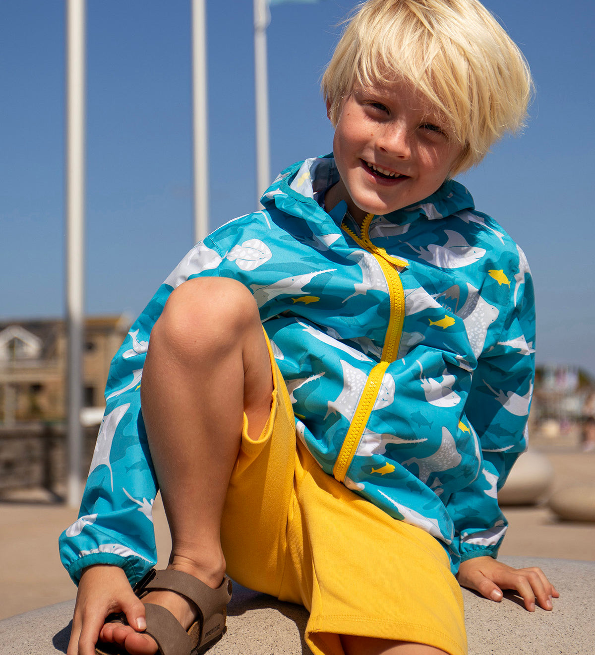 Child smiling and sitting on a concrete surface at the beach. Wearing the light blue hooded Frugi rain jacket with a repeating grey manta ray and shark print