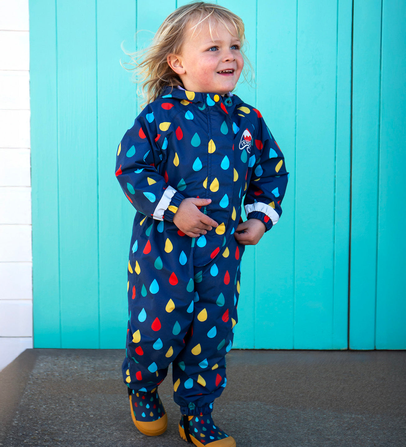 Child standing on a concrete surface in front of a white and turquoise beach hut. Wearing a navy Frugi all in one waterproof hooded suit with a repeating coloured raindrop print and reflective strips on each arm. 