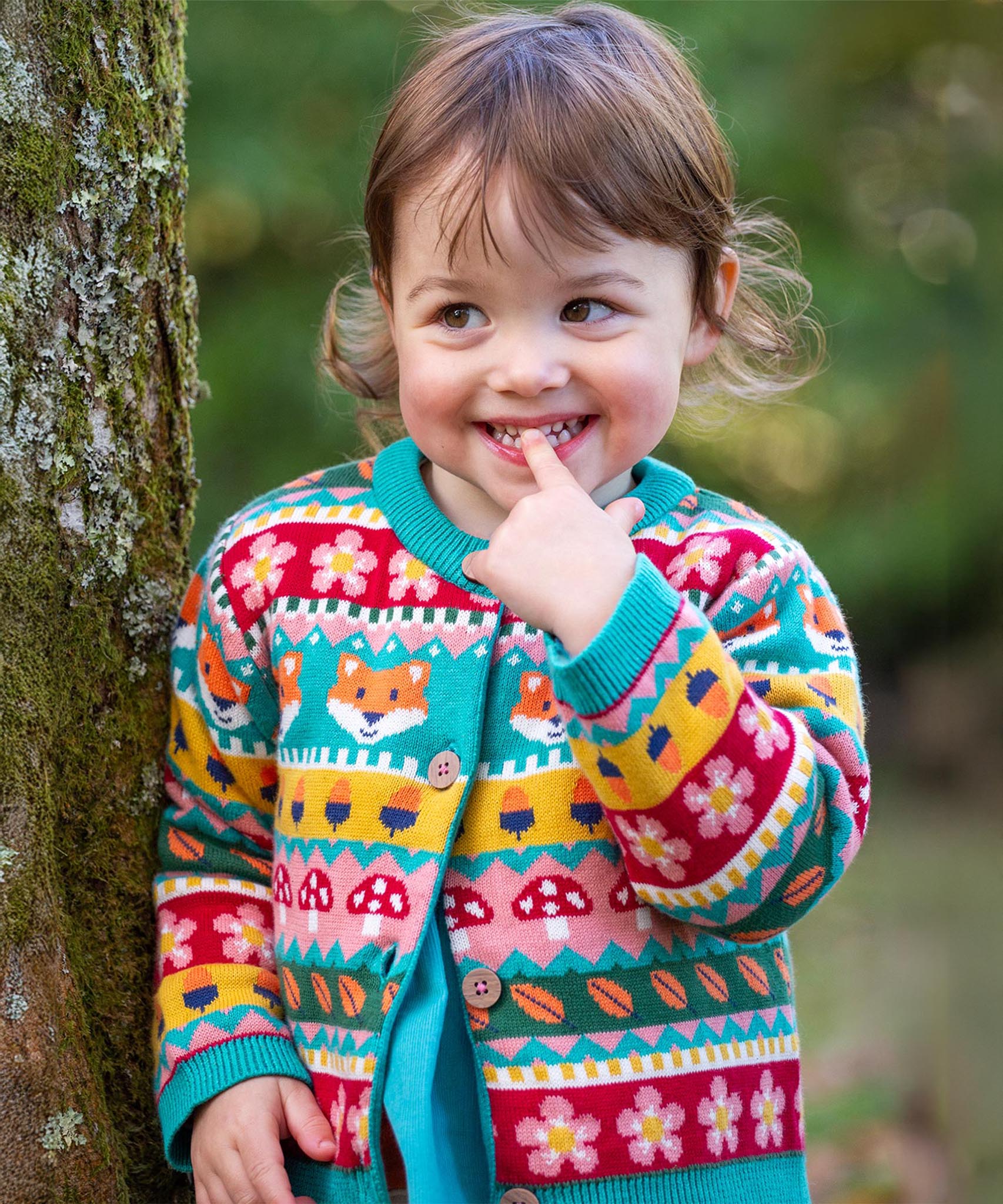 A child leaning on a tree and wearing the Frugi kids robyn fairisle cardigan in fox. A 100% GOTS organic cotton cardigan with a bright woodland fairisle design. 