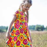 A child smiling and walking through a grassy field. Wearing the yellow Frugi skater dress with a repeating pink and orange floral print