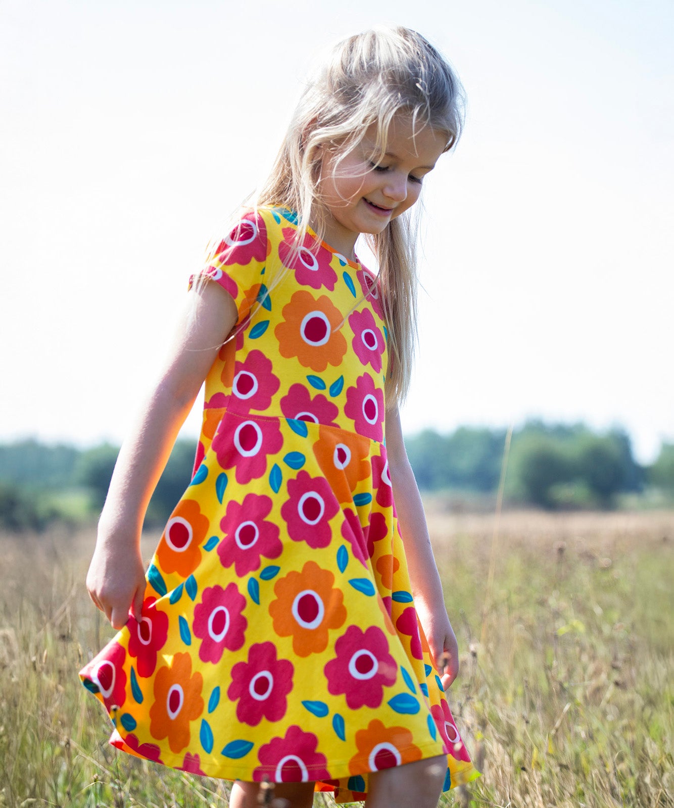 A child smiling and walking through a grassy field. Wearing the yellow Frugi skater dress with a repeating pink and orange floral print