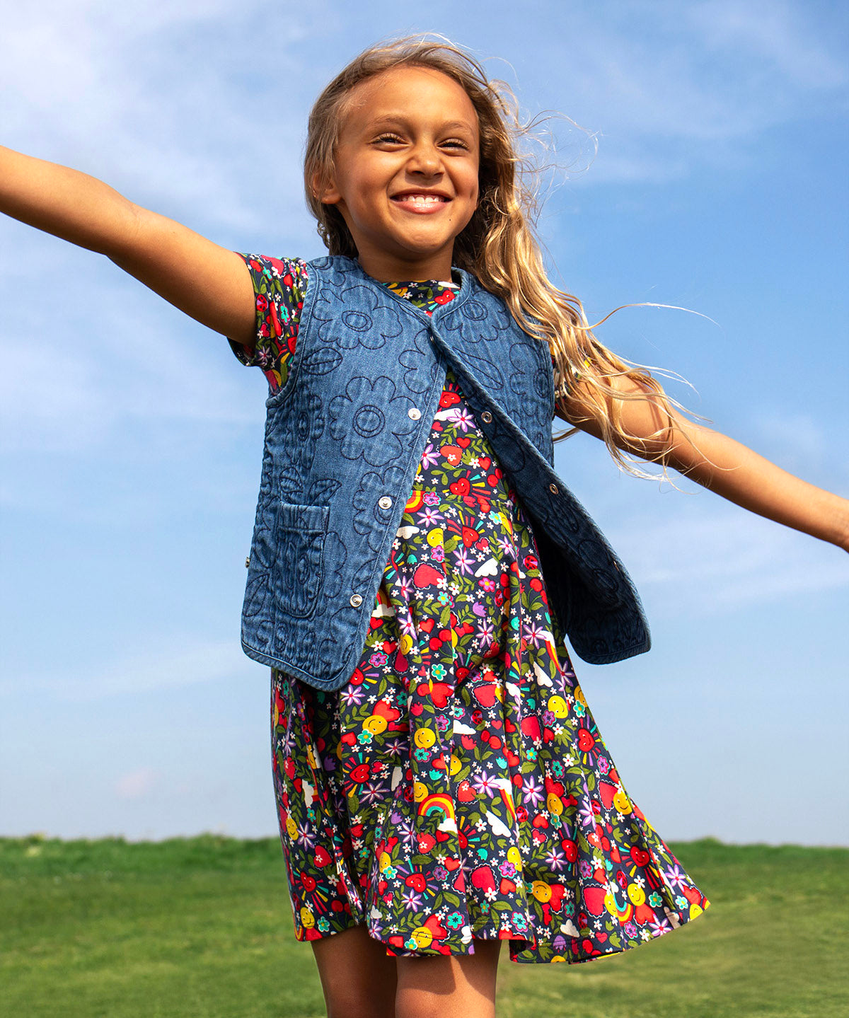 A child smiling and standing outdoors with a grassy hill in the backgroud. Wearing the navy Frugi organic cotton skater dress. In navy with a repeating heart, sun rainbow and flower print with a denim gilet