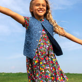 A child smiling and standing outdoors with a grassy hill in the backgroud. Wearing the navy Frugi organic cotton skater dress. In navy with a repeating heart, sun rainbow and flower print with a denim gilet
