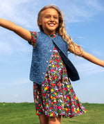 A child smiling and standing outdoors with a grassy hill in the backgroud. Wearing the navy Frugi organic cotton skater dress. In navy with a repeating heart, sun rainbow and flower print with a denim gilet