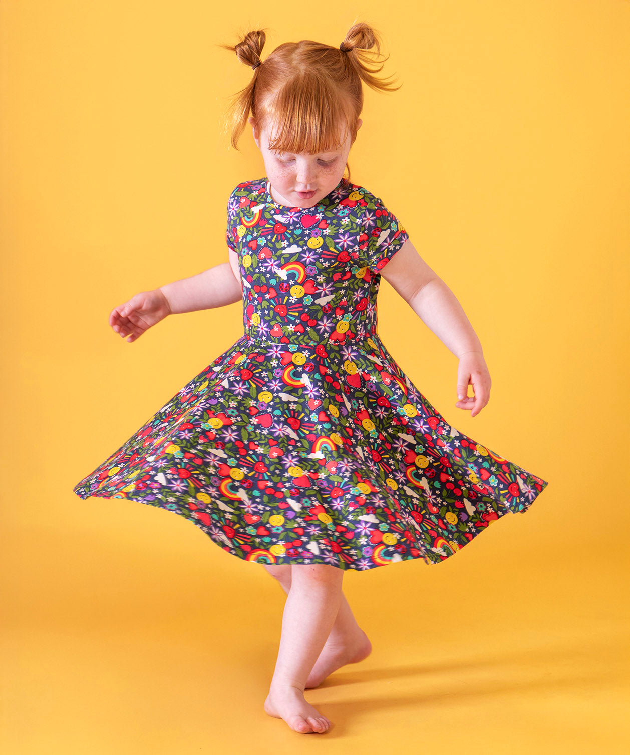 A child doing ballet in front of a light orange background. Wearing the Frugi organic cotton skater dress. In navy with a repeating heart, sun rainbow and flower print.