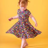 A child doing ballet in front of a light orange background. Wearing the Frugi organic cotton skater dress. In navy with a repeating heart, sun rainbow and flower print.