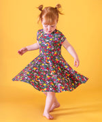 A child doing ballet in front of a light orange background. Wearing the Frugi organic cotton skater dress. In navy with a repeating heart, sun rainbow and flower print.