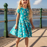 A child smiling and standing on a concrete surface at the seaside. Wearing the Frugi skater dress. In light blue with a repeating grey manta ray and rainbow print