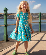 A child smiling and standing on a concrete surface at the seaside. Wearing the Frugi skater dress. In light blue with a repeating grey manta ray and rainbow print