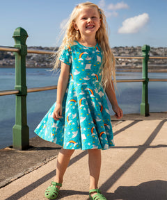 A child smiling and standing on a concrete surface at the seaside. Wearing the Frugi skater dress. In light blue with a repeating grey manta ray and rainbow print