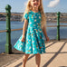 A child smiling and standing on a concrete surface at the seaside. Wearing the Frugi skater dress. In light blue with a repeating grey manta ray and rainbow print