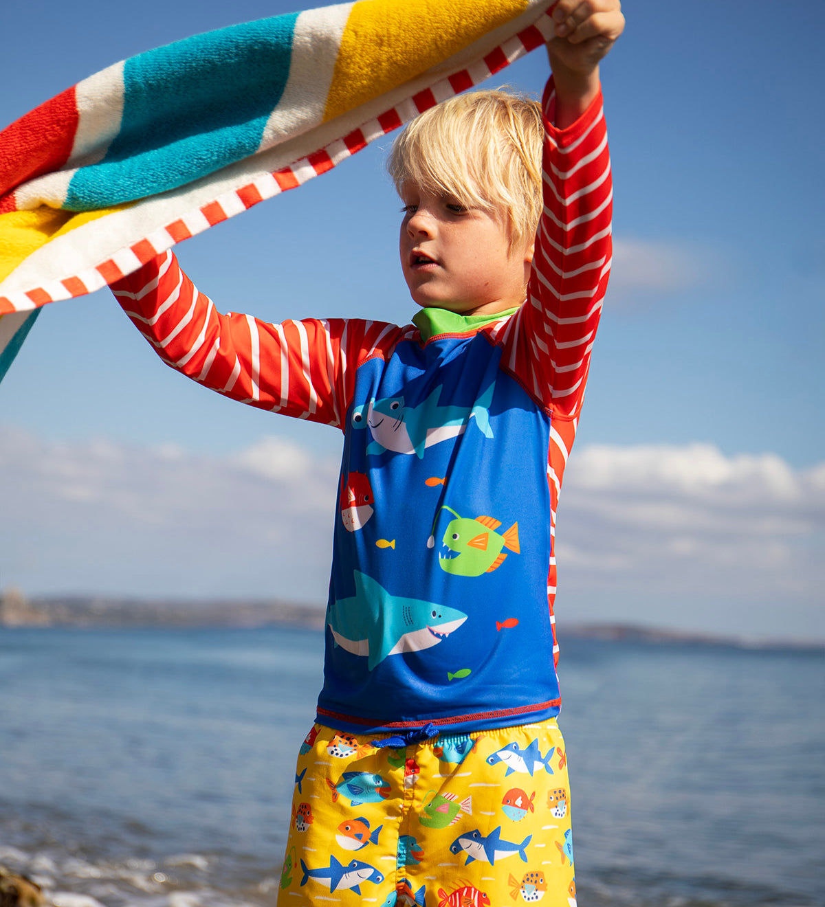 Child standing on the beach with the ocean in the background. Holding a striped beach towel and wearing a blue rash vest with red and white stripes and yellow swim shorts