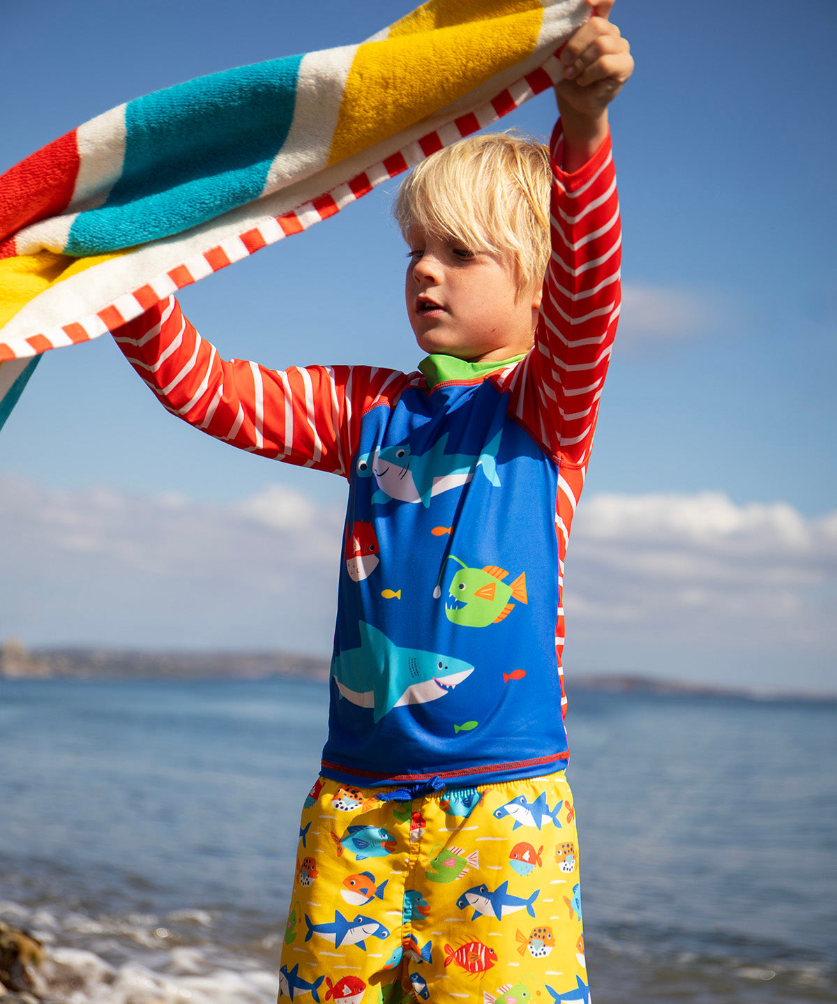 Child standing on the beach with the ocean in the background. Holding a striped beach towel and wearing a blue rash vest with red and white stripes and yellow swim shorts