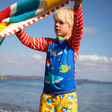 Child standing on the beach with the ocean in the background. Holding a striped beach towel and wearing a blue rash vest with red and white stripes and yellow swim shorts
