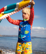Child standing on the beach with the ocean in the background. Holding a striped beach towel and wearing a blue rash vest with red and white stripes and yellow swim shorts