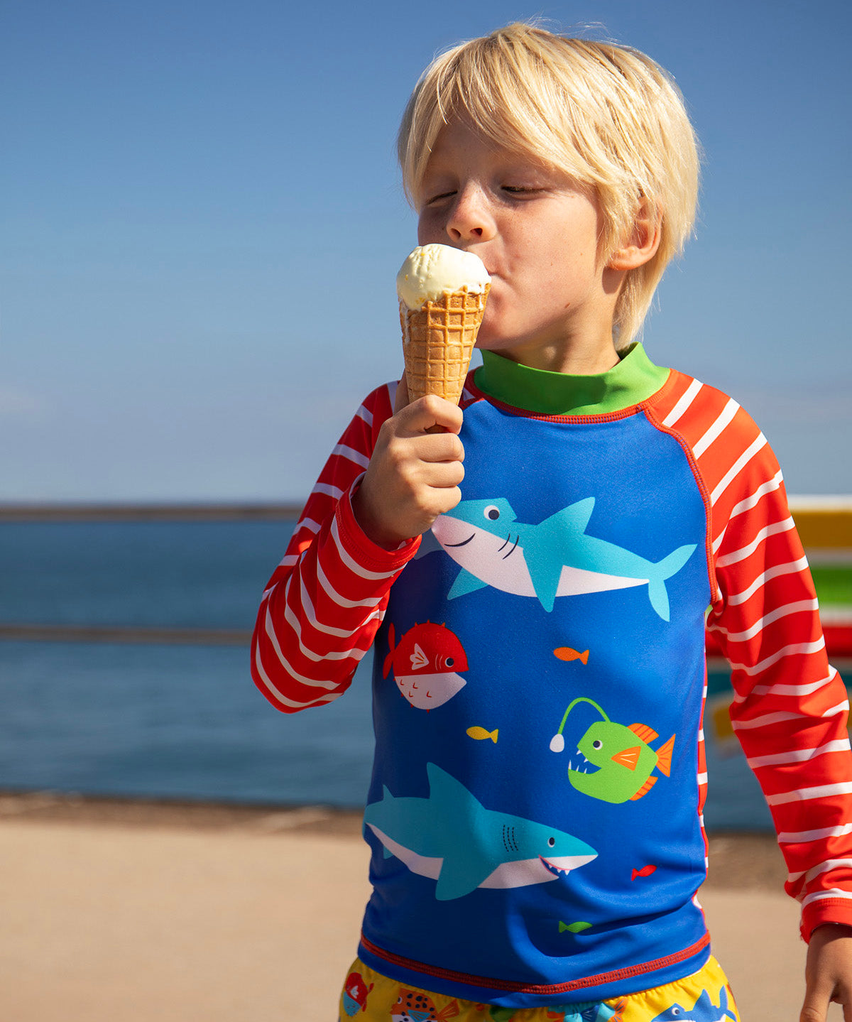 Child standing on the beach with the ocean in the background and holding a i cream. Wearing a blue rash vest with red and white stripes 