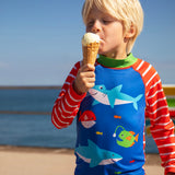 Child standing on the beach with the ocean in the background and holding a i cream. Wearing a blue rash vest with red and white stripes 